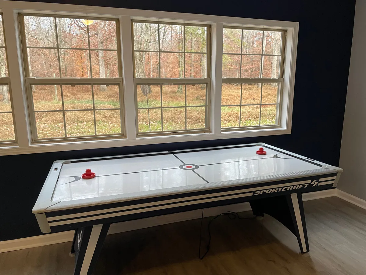 The living space features an air hockey table set near large windows with a view of lush greenery outside. Friends are shown playing air hockey, capturing the fun, social nature of the room. The abundance of natural light and vibrant activity create an inviting and playful mood.