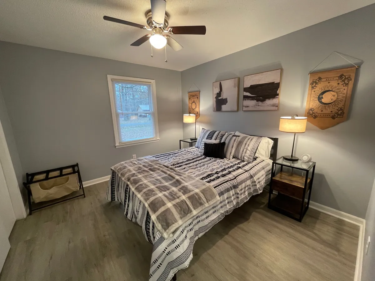 A cozy bedroom showcasing a comfortable bed with patterned bedding, soft lighting from bedside lamps, and a window letting in natural light. Framed artwork adds personality, while the window connects the space to the outdoors, suggesting a peaceful environment for rest.