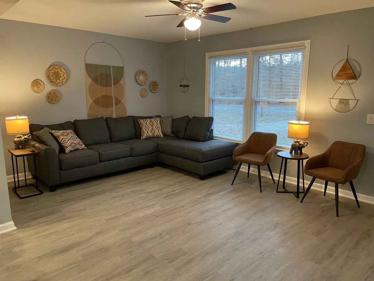 This image showcases a cozy sunroom interior, highlighted by a comfortable sectional sofa, decorative wall art, table lamps, and large windows that let in abundant natural light. The layout encourages both relaxation and socializing, making it an ideal setting for reading, working, or spending time with friends and family.