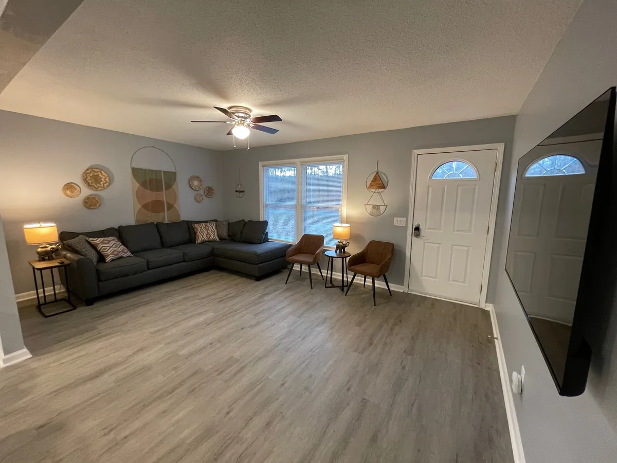 This image showcases a sectional sofa arranged invitingly in the main living room, complemented by decorative wall art, ample windows, an entry door, and table lamps. The setting exudes a cozy atmosphere ideal for relaxing with family or friends, watching TV, reading, or entertaining guests.