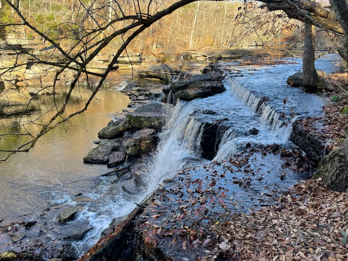 A picturesque waterfall cascades over rugged rock formations, surrounded by vibrant autumn foliage. The calm water at the base of the falls reflects the serene landscape, and the scene is inviting for nature walks, photography, or simply relaxing by the water. The natural ambiance is enhanced by the soothing sound of the falls and the changing colors of the leaves.