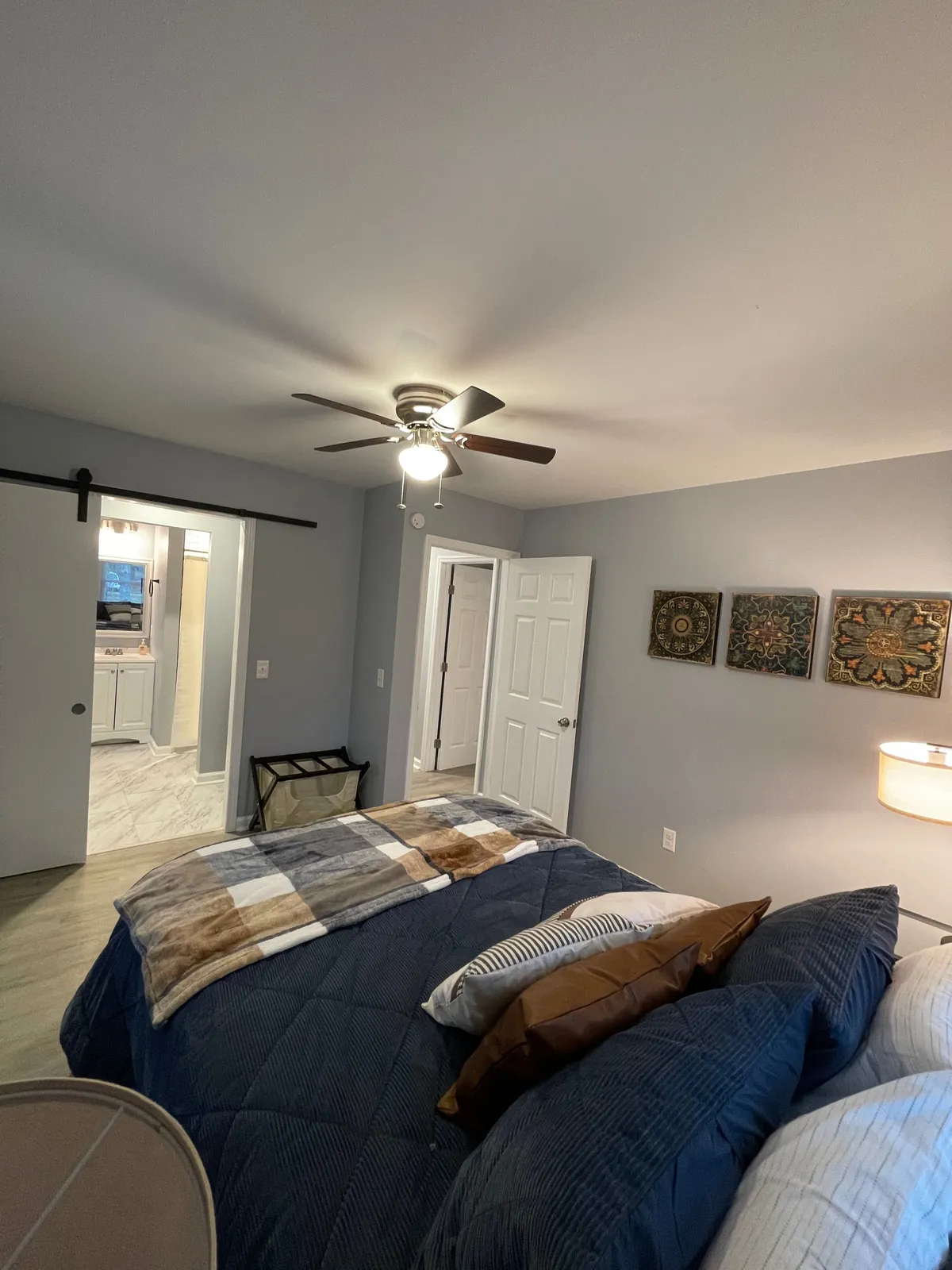 This interior shot features a bedroom with a bed draped in patterned throw blankets, decorative wall art, and a ceiling fan overhead. Sunlight streams into the room, creating a warm and inviting mood for both restful moments and quiet reading.